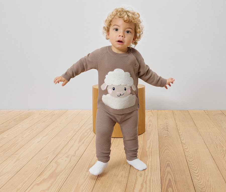 Egy göndör hajú kisgyermek áll egy zsámolyon, barna kötött pulóvert és leggingset visel birka mintával.