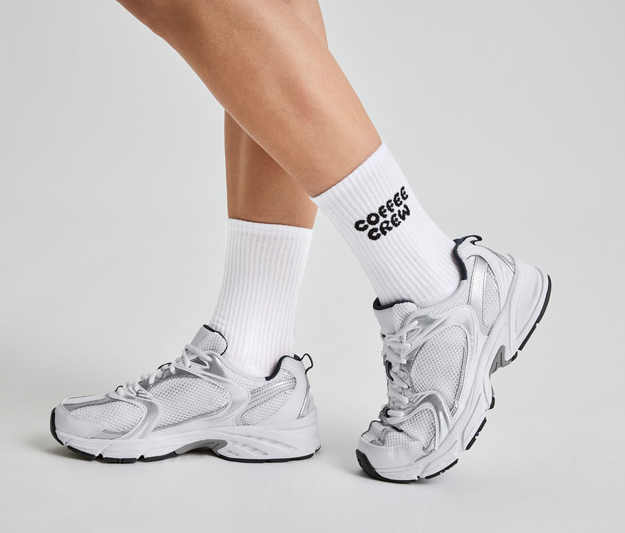 Egy személy lábai "Coffee Crew" feliratú fehér zokniban, valamint fehér és ezüst sportcipőben.