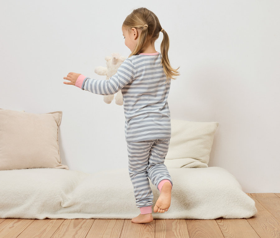 Kislány szürke-fehér csíkos pizsamában plüssmackót tart a kanapén.