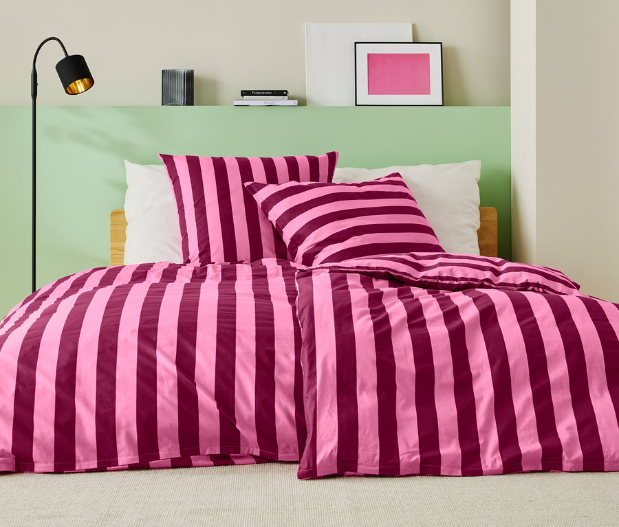 Ágy rózsaszín-bordó csíkos ágyneművel, két párnával és egy állólámpával.
