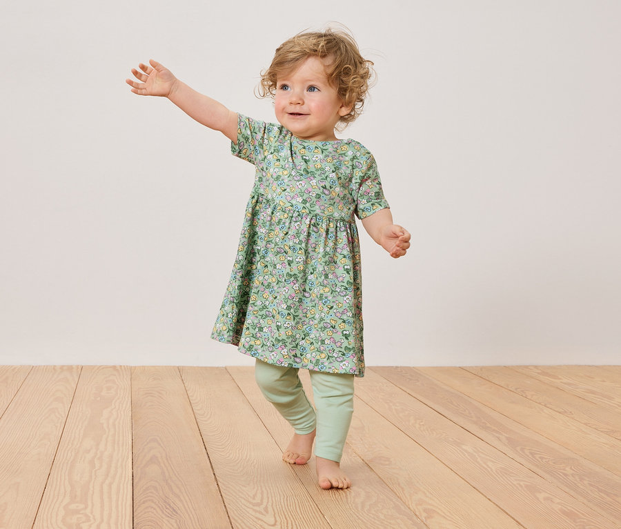 Egy szőke fürtös kisgyermek virágos ruhát és zöld leggingset visel, miközben egy fapadlón áll.