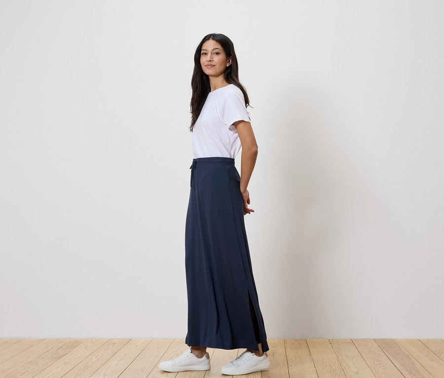 Nő áll oldalt, fehér pólót, hosszú, sötétkék szoknyát és fehér tornacipőt visel.