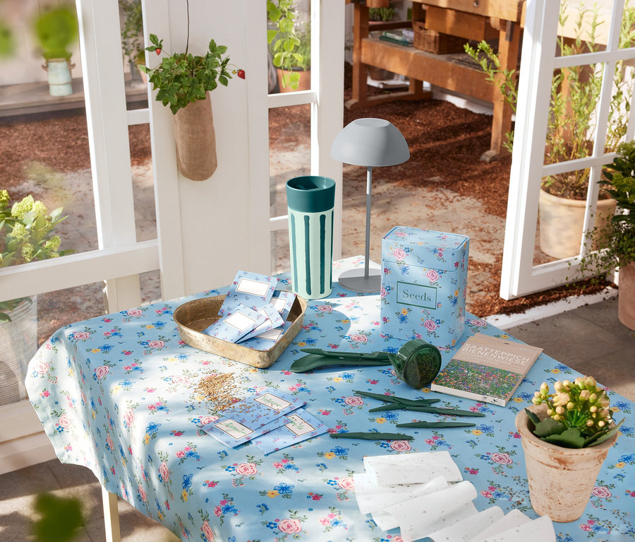 Egy szürke LED akkumulátoros asztali lámpa áll egy virágos terítővel borított asztalon, kerti eszközök és magvak mellett.