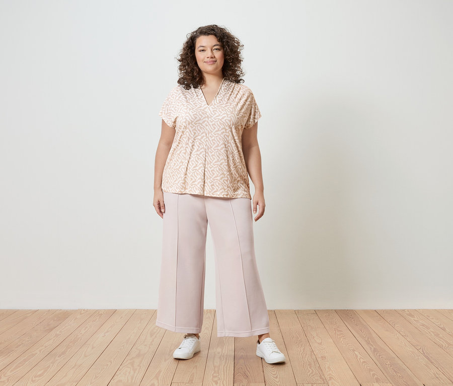 Egy göndör hajú nő világos rózsaszín mintás blúzt, rózsaszín culotte nadrágot és fehér tornacipőt visel egy fehér fal előtt.