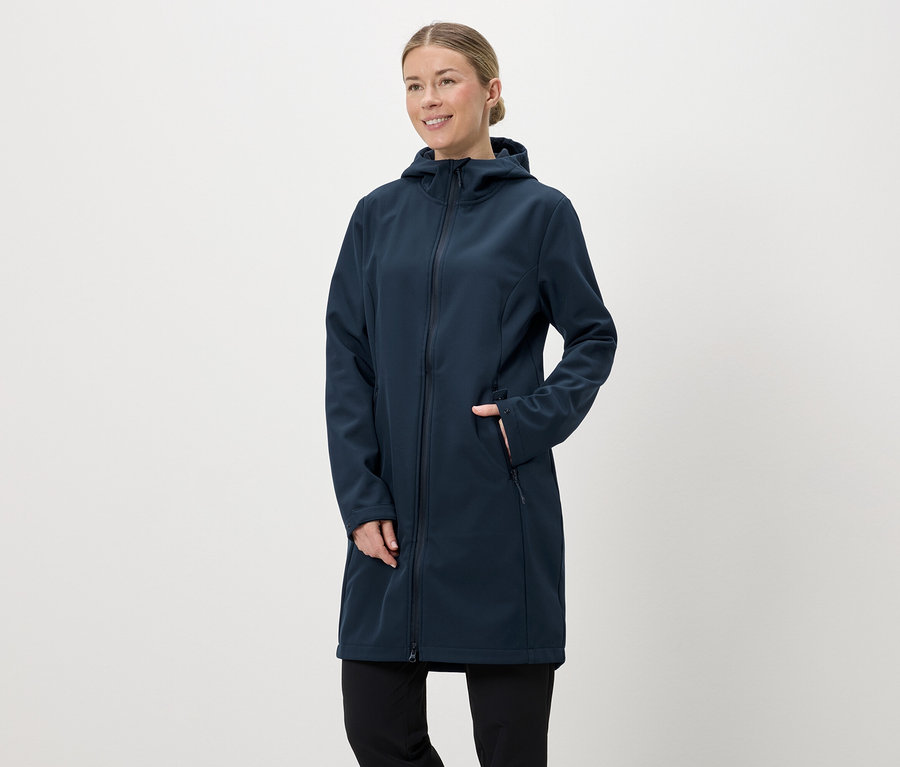 Nő sötétkék, hosszú softshell kabátban, kapucnival és cipzárral, kezét a zsebébe teszi.