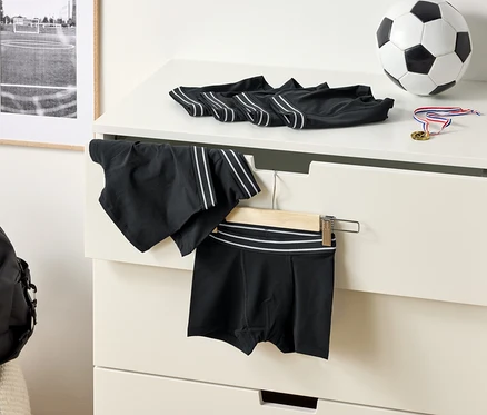 A komódon fekete, fehér csíkos boxeralsók, egy focilabda és egy érem látható. Egy sport rövidnadrág lóg egy vállfán.