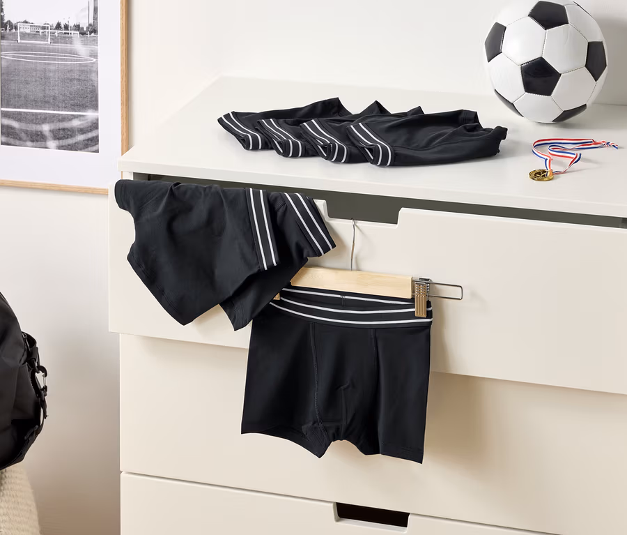 A komódon fekete, fehér csíkos boxeralsók, egy focilabda és egy érem látható. Egy sport rövidnadrág lóg egy vállfán.
