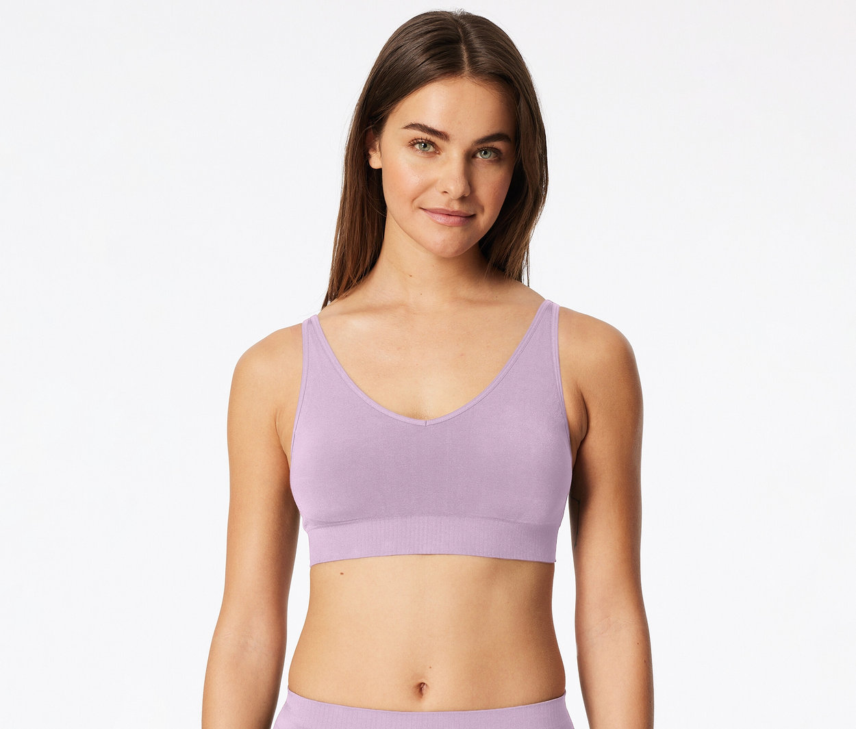 Hosszú barna hajú nő világoslila V-nyakú, széles pántos bralette-et visel, előre néz fehér háttér előtt.