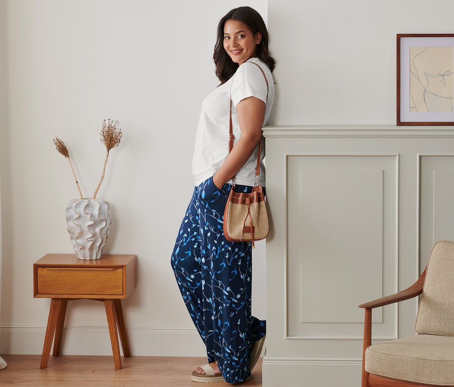 Egy nő a falnak támaszkodva pózol virágos, bőszárú nadrágban, fehér len felsőben és bézs bőr szandálban. Egy barna fonott oldaltáska van a vállán.