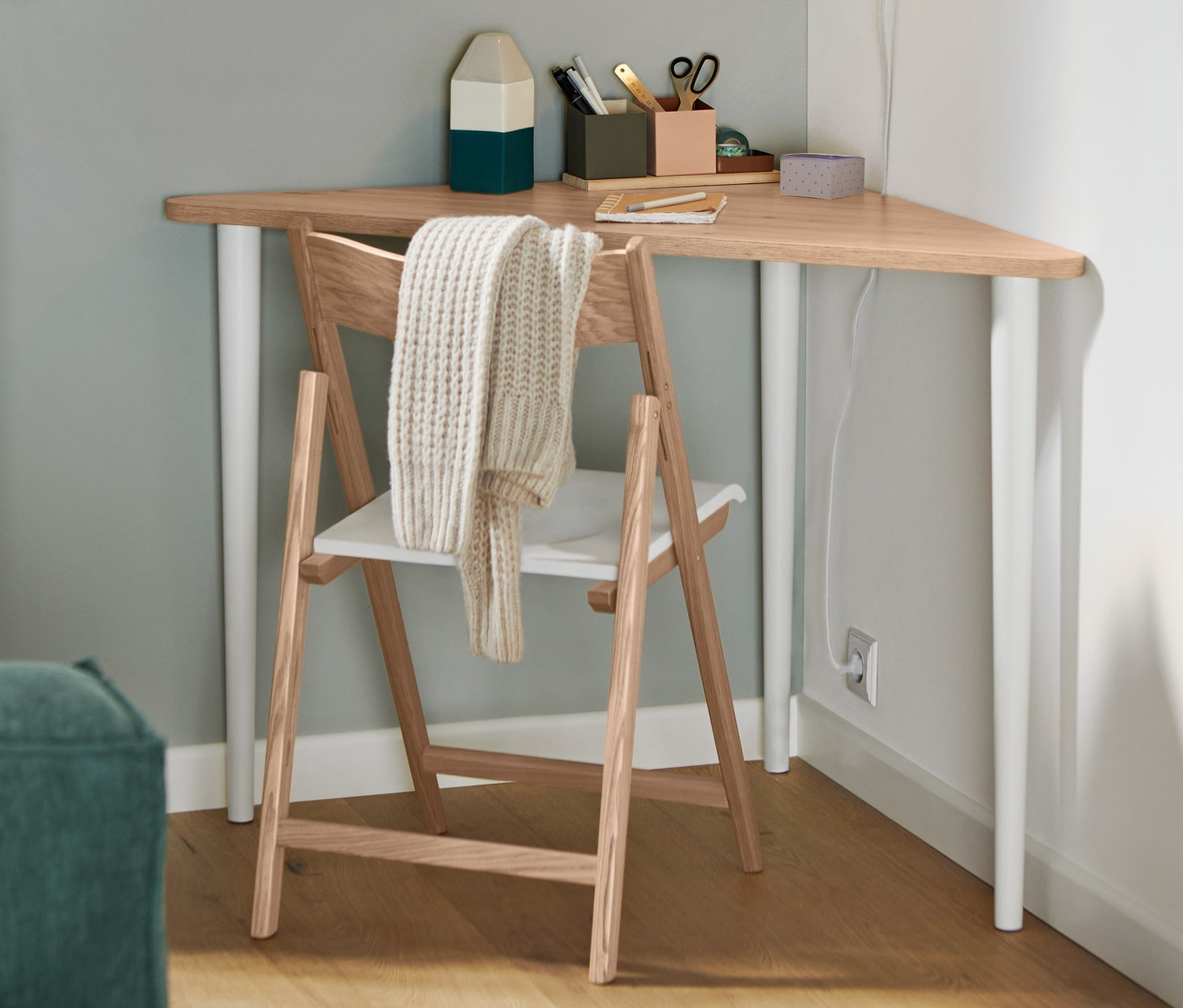 Egy összecsukható, FSC® tanúsítvánnyal rendelkező, kőrisfából készült szék áll egy háromszög alakú asztalnál. A falon egy fehér fém fali lámpa.