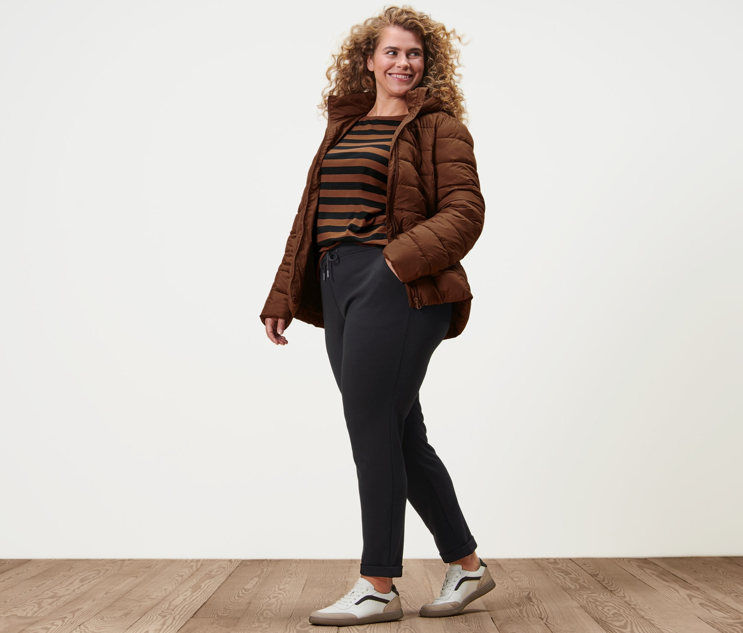 Egy nő sétál, és konyak színű, kapucnis steppelt kabátot, 3/4-es ujjú, csíkos felsőt, női lezser nadrágot és bőr sneaker cipőt visel.