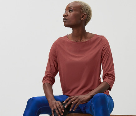 Nő ül a padon, és egy vizes palackot tart. Terrakotta színű, 3/4-es ujjú funkcionális felsőt visel.