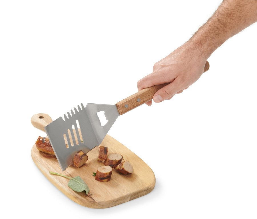 Egy kéz egy multifunkciós grillező eszközt tart egy fából készült vágódeszka fölött, amin felvágott kolbász van.