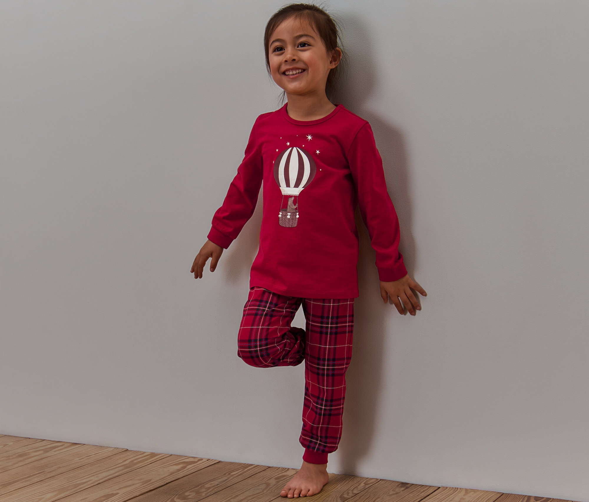 Egy mosolygós kislány áll a fal mellett, piros pizsamában, kockás nadrággal és egy világító léggömb mintával.