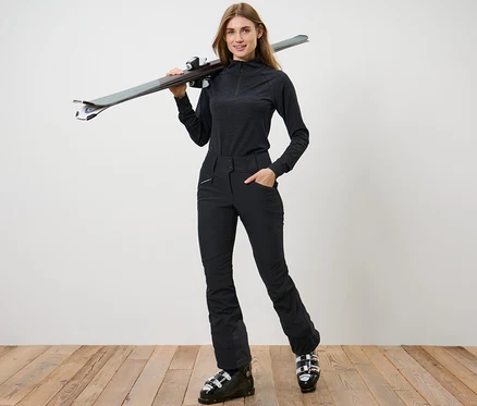 Nő pózol fekete női softshell jet sínadrágban és síbakancsban, a vállán sílécekkel.