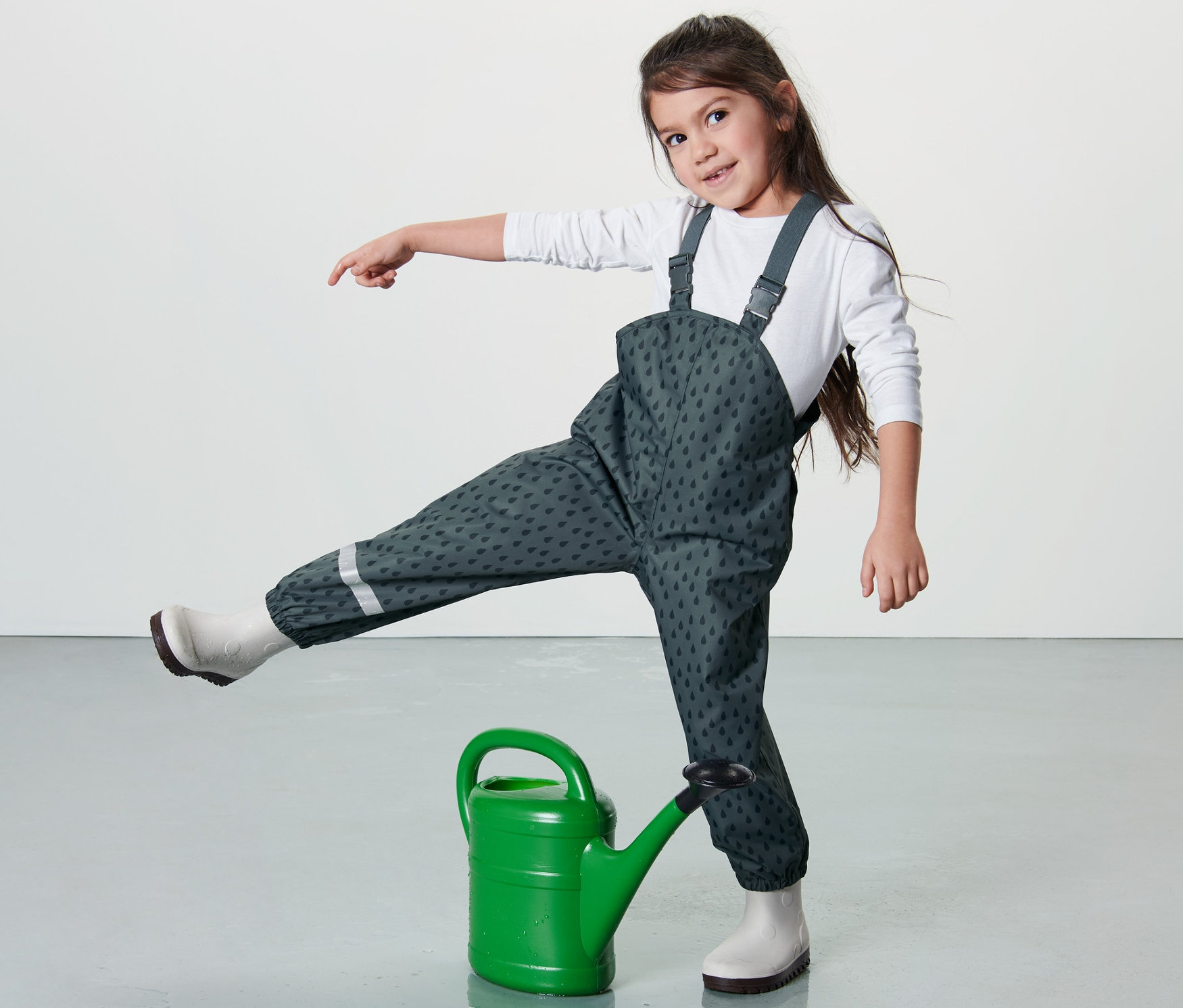 Egy kislány az egyik lábával egy zöld locsolókannán áll, és palakék kisgyerek kantáros esőnadrágot visel.