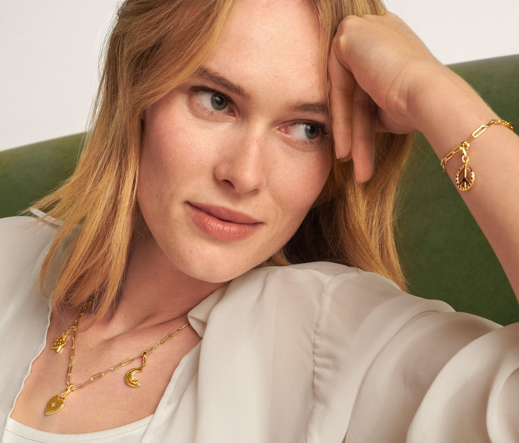 Egy szőke nő ül egy zöld kanapén, és egy arany Nyaklánc Collect, egy Karkötő Collect, egy Függőszett Love and Peace és egy Függőszett, Moon and Stars szettet visel.