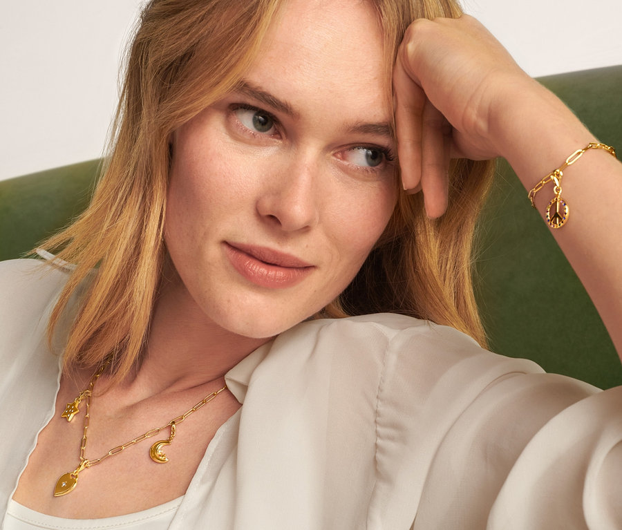 Egy szőke nő ül egy zöld kanapén, és egy arany Nyaklánc Collect, egy Karkötő Collect, egy Függőszett Love and Peace és egy Függőszett, Moon and Stars szettet visel.