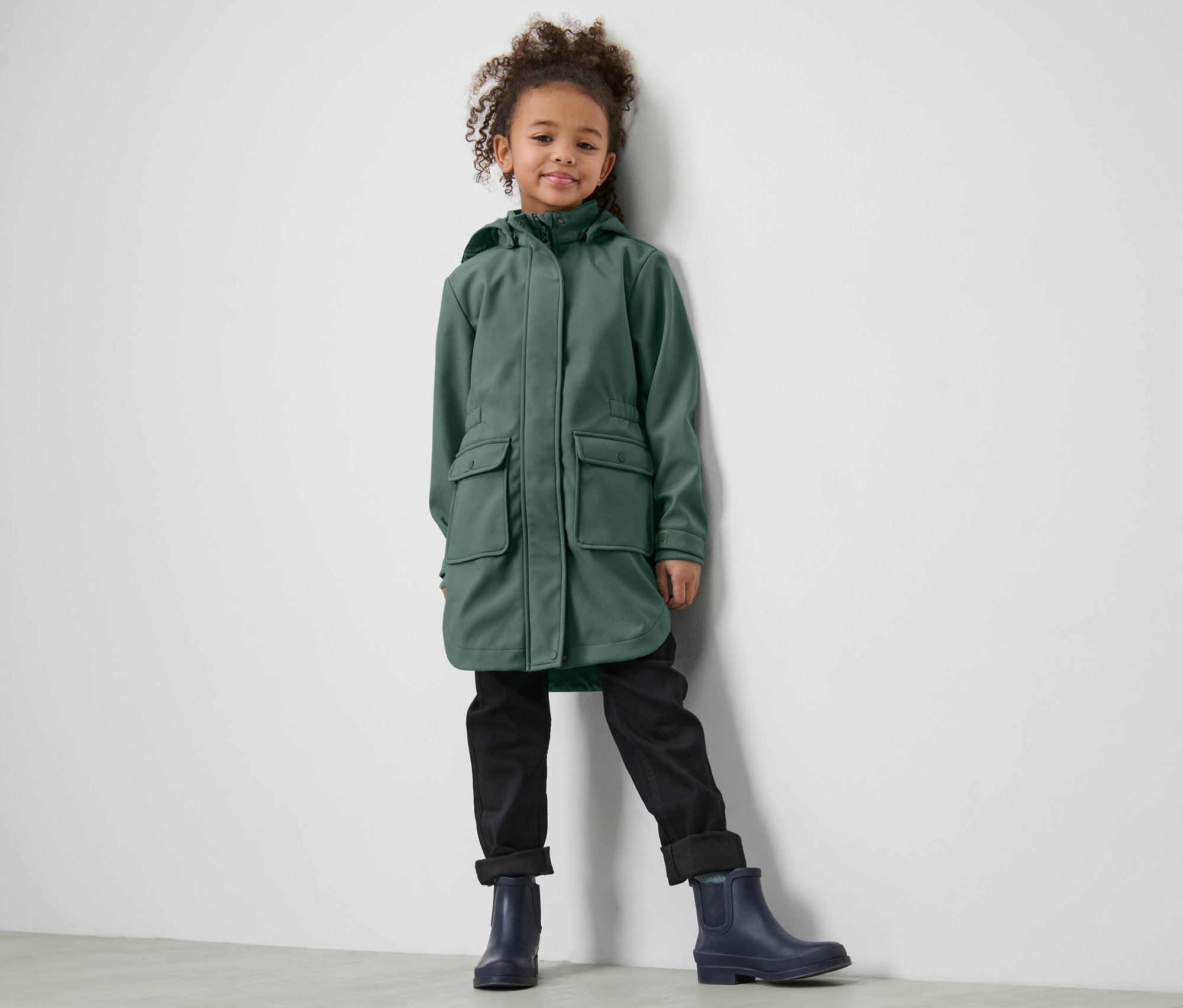 Lány áll a falnak támaszkodva gyerek softshell kabátban, fekete nadrágban és kék cipőben.