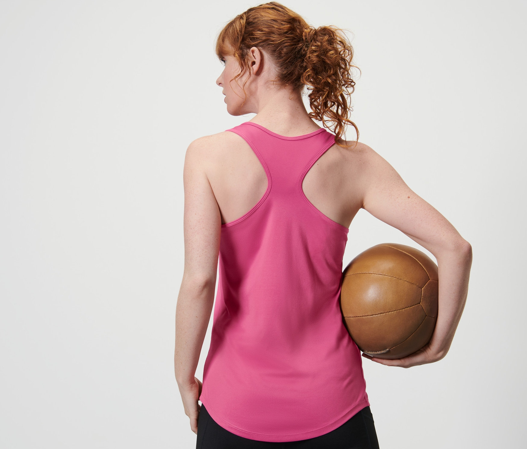 Nő hátulról tart egy barna labdát egy rózsaszín funkcionális sporttopban egy kétrészes szettből és fekete leggingsben.
