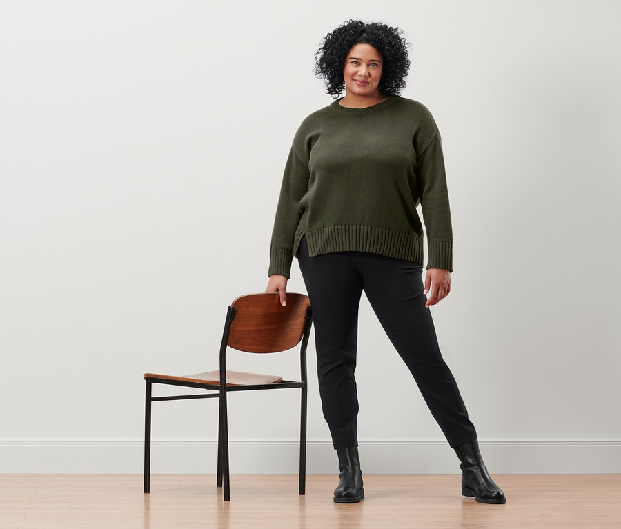 Nő pózol egy szék mellett, fekete sztreccsnadrágot és olíva kötött pulóvert visel.