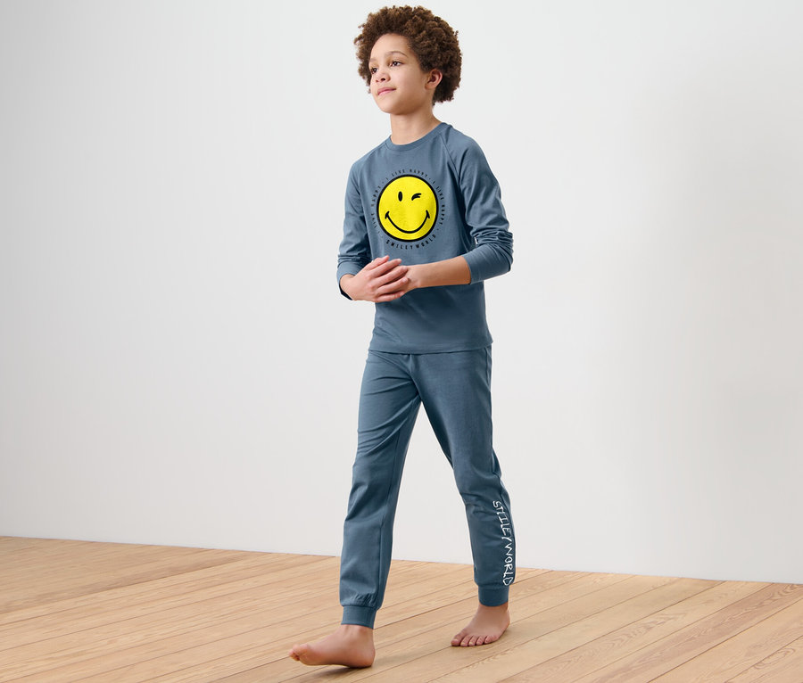 Fiú kék, hosszú ujjú pizsamában, sárga "Smiley" mosolygóval a mellkasán és "Smiley" nadrággal, mezítláb áll a fapadlón. 2 fiú pizsama, Smiley.
