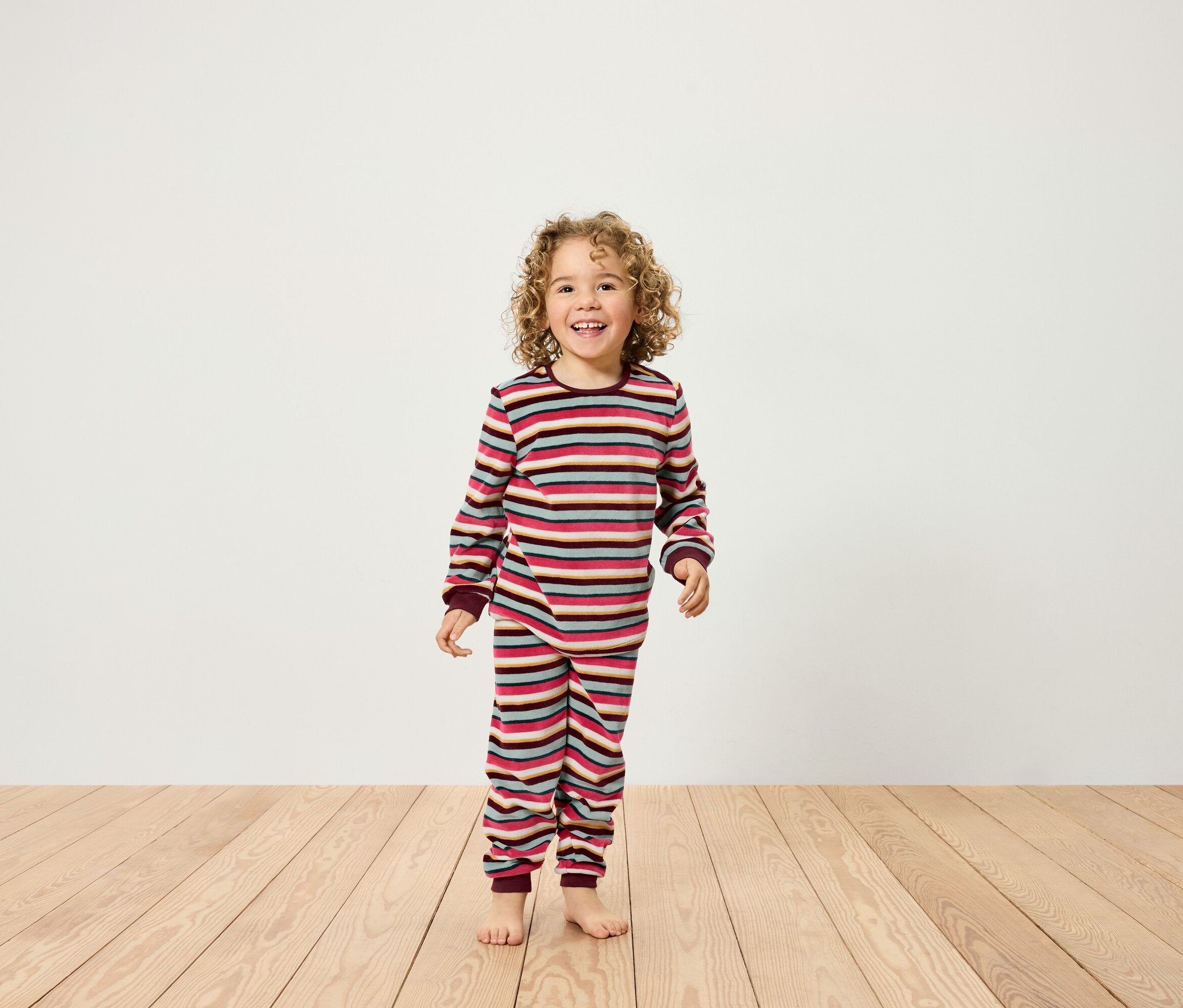 Egy mosolygó, göndör hajú gyermek áll egy fapadlón, csíkos pizsamában.