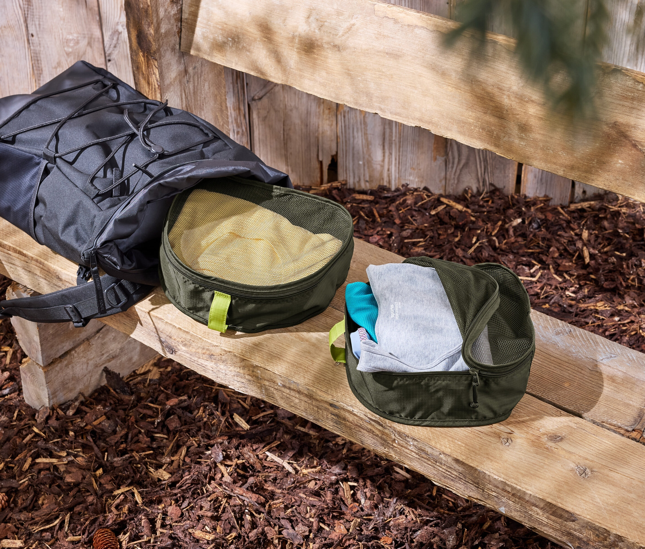 Egy fekete hátizsák fekszik egy fa padon két zöld ruhaszervező mellett.