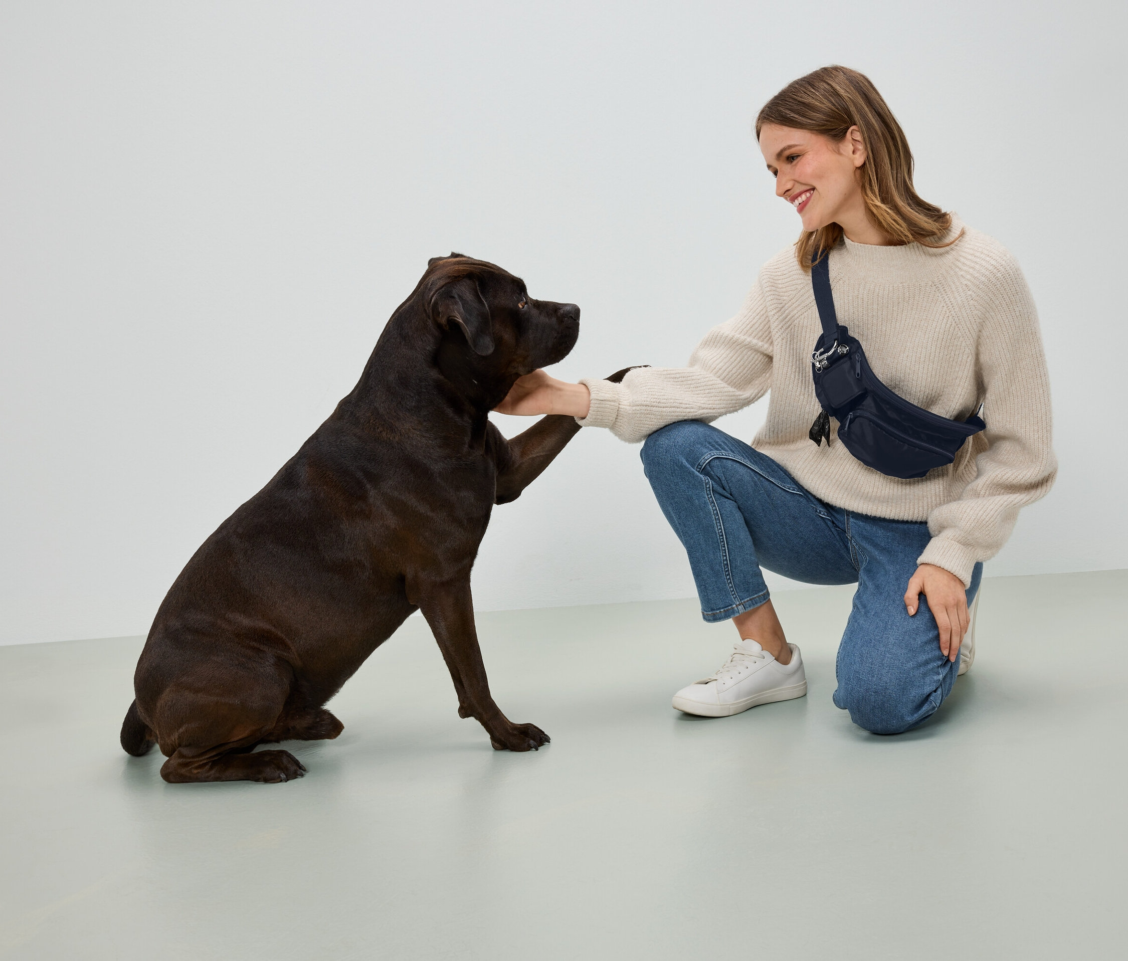 Egy bézs pulóvert és kék farmert viselő nő térdelve fogja egy sötétbarna kutya mancsát. Sötétkék övtáskát visel.