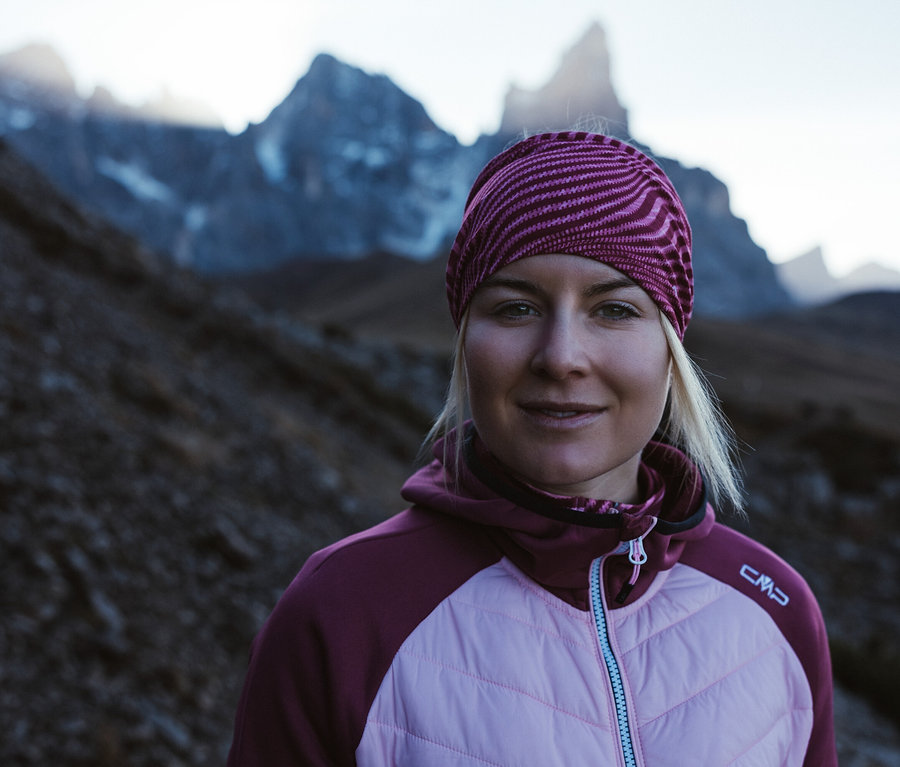 Szőke nő, csíkos fejpánttal, beere színű CMP női hibrid dzsekiben, hegyvidéki háttér előtt.