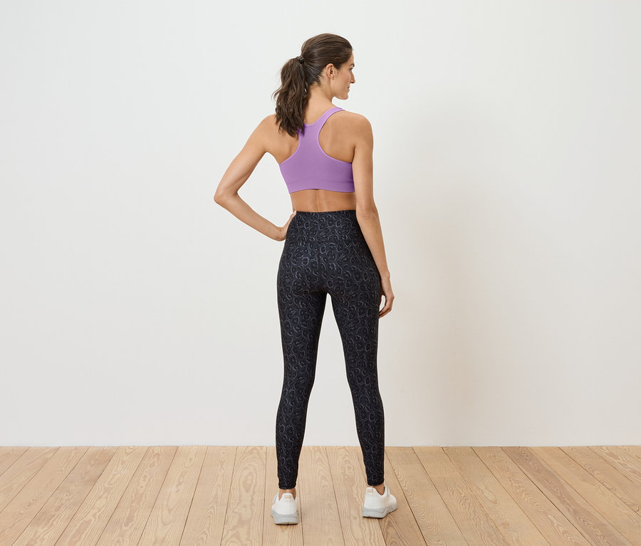 Egy nő háttal áll, lila sportmelltartót és fekete leggingset visel.