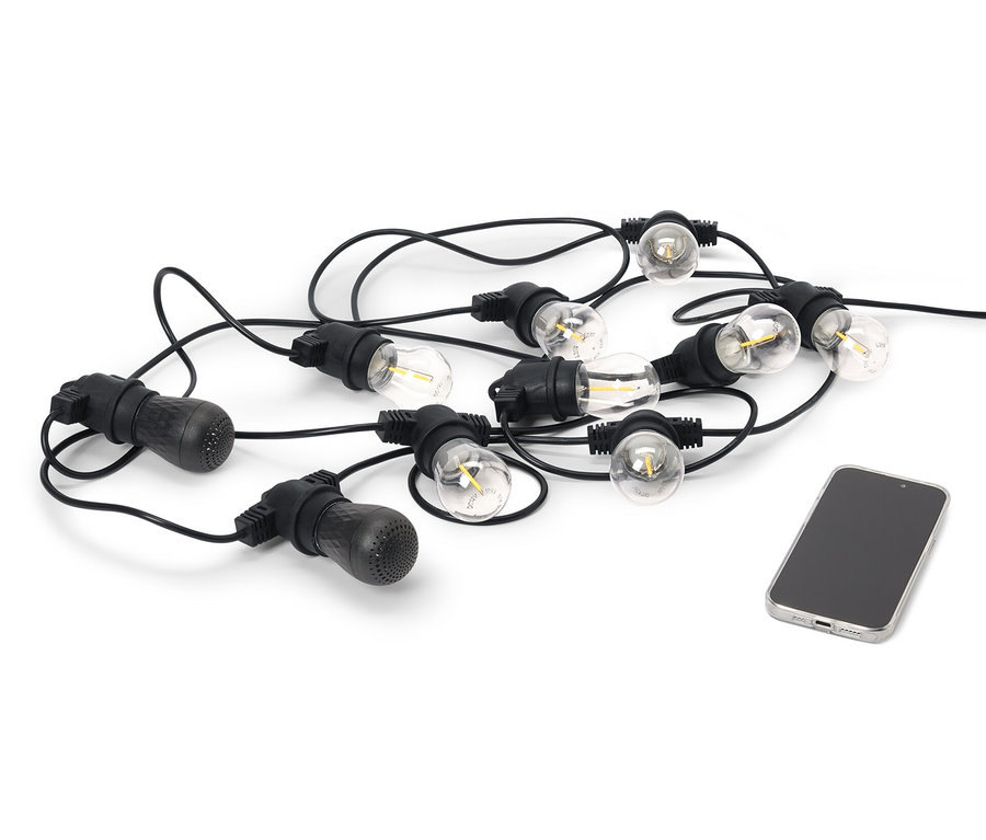 Egy fekete kültéri LED-es fényfüzér 2 Bluetooth® hangszóróval egy mobiltelefon mellett fekszik fehér háttéren.