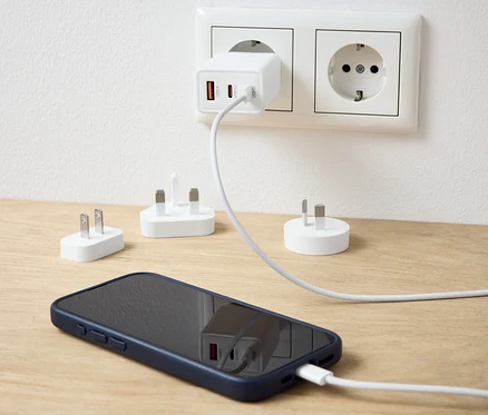 Egy fehér USB adapter van a konnektorba dugva, és egy mobiltelefont tölt. Az asztalon további adapterek találhatók.