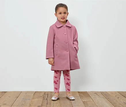 Egy lány áll egy fehér fal előtt, rózsaszín kabátot, csillagos leggingset és ezüst cipőt visel.
