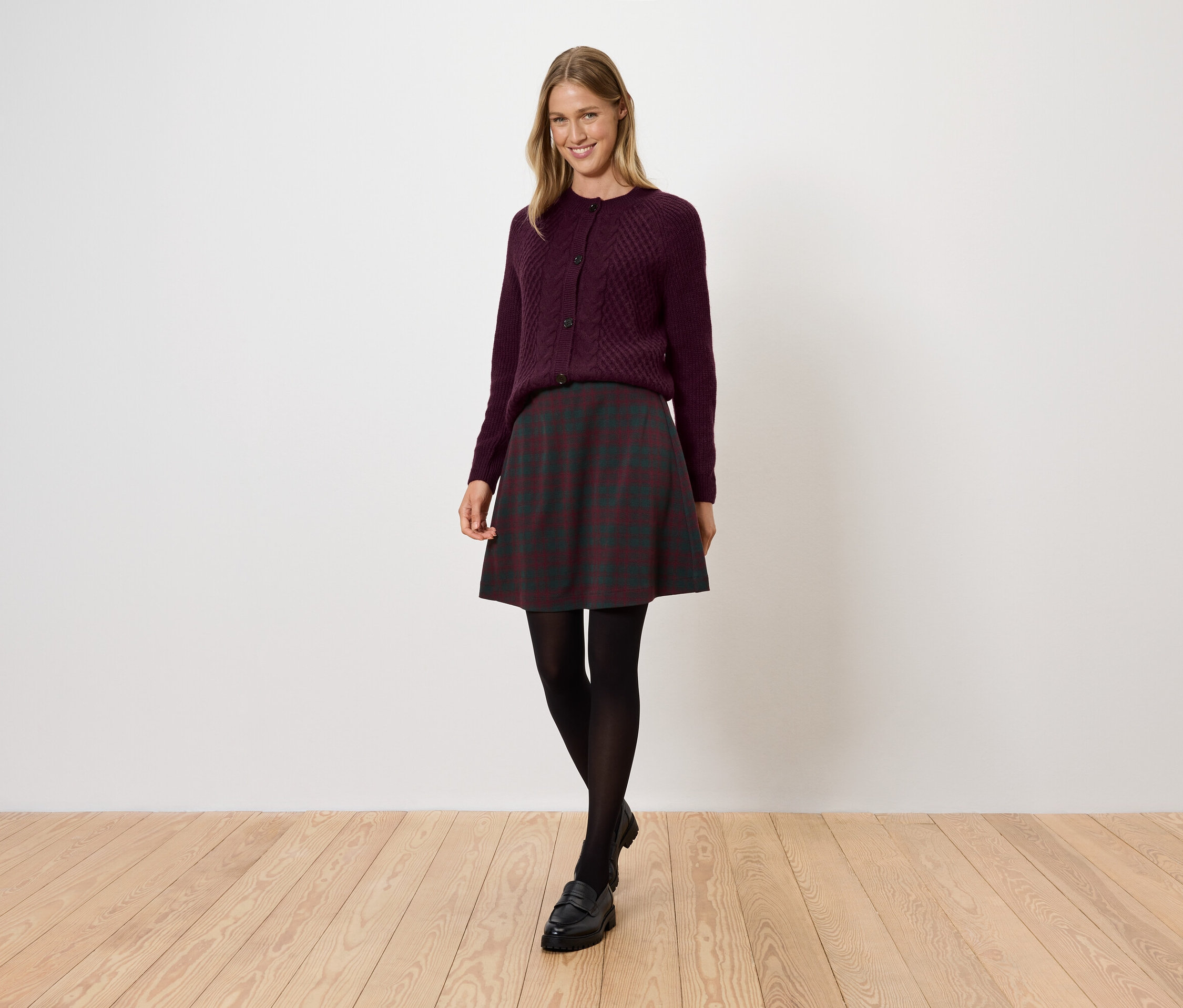 Nő bordó pulóverben, sötét szoknyában, fekete harisnyában és cipőben áll a fapadlón.