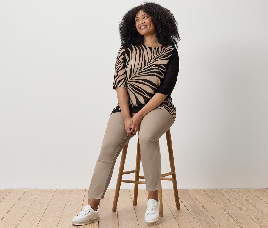 Nő ül egy széken, 7/8-os tregginget, mintás felsőt és tornacipőt visel.