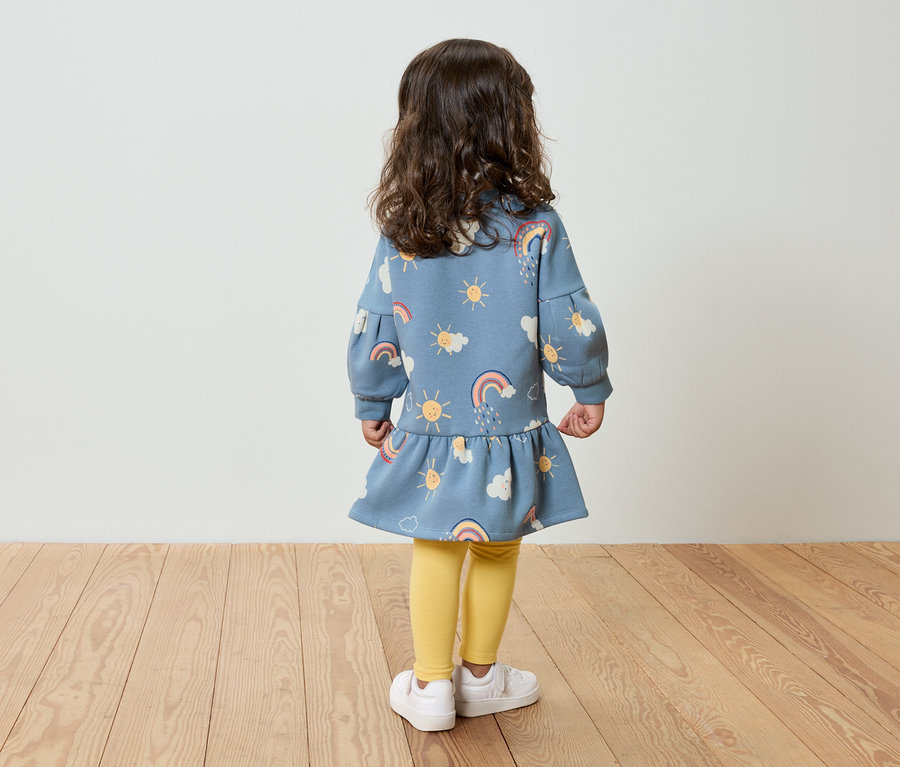 Egy kislány áll háttal a kamerának, kék ruhát visel, amelyen nap, felhő és szivárvány minta van, sárga leggings és fehér cipő van rajta.