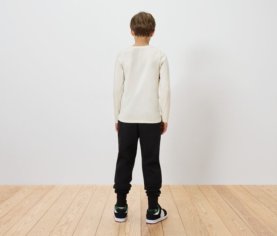 Egy gyermek háttal áll a nézőnek, bézs hosszú ujjú inget, fekete nadrágot és zöld-fekete tornacipőt visel.