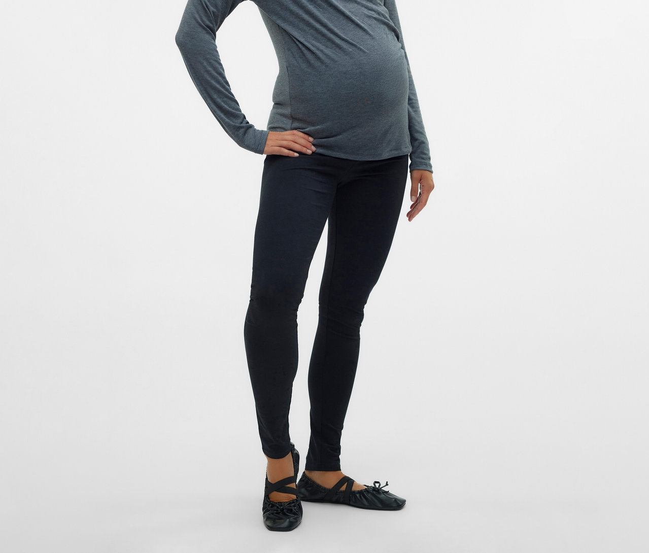 Várandós nő szürke felsőben és 2 db MAMALICIOUS kismama leggingsben, fekete színben, csípőre tett kézzel pózol fehér háttér előtt.