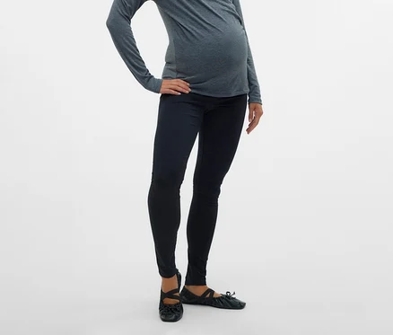 Várandós nő szürke felsőben és 2 db MAMALICIOUS kismama leggingsben, fekete színben, csípőre tett kézzel pózol fehér háttér előtt.