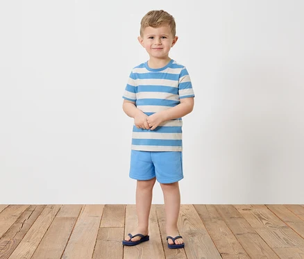 Egy szőke hajú fiú mezítláb áll egy fapadlón, kék-fehér csíkos pólót, kék rövidnadrágot és kék flip-flop papucsot visel.