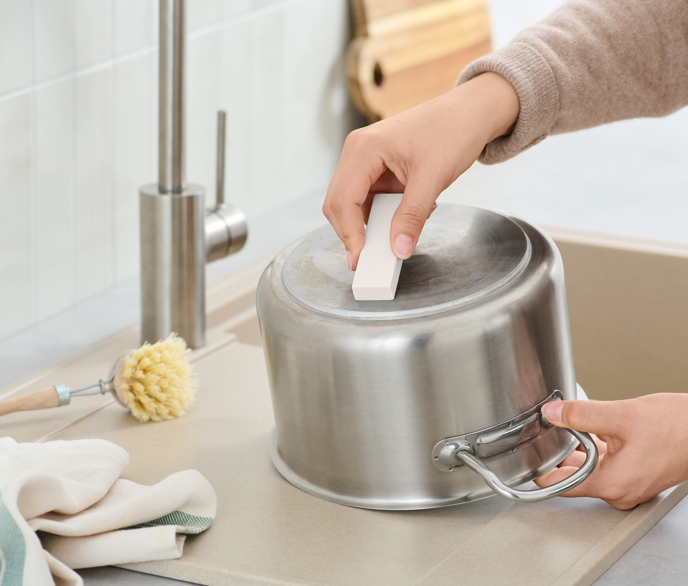 Egy személy tisztít egy rozsdamentes acél edényt egy tisztítószivaccsal a mosogatóban, egy kefe és egy rongy mellett.