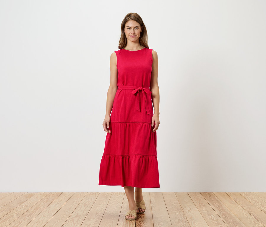 Egy nő áll egy fehér fal előtt, piros, ujjatlan, fodros ruhát visel övvel és szandállal.