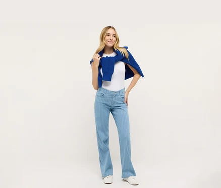 Nő pózol fehér pólóban és kék Angels denim »Liz« bőszárú farmernadrágban, kék pulóverrel a vállán.