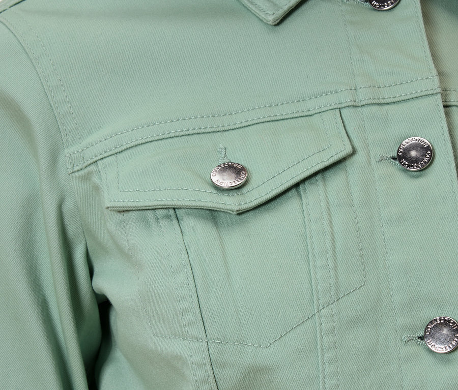 Egy világoszöld dzseki közeli képe ezüst gombokkal és egy mellzsebbel.