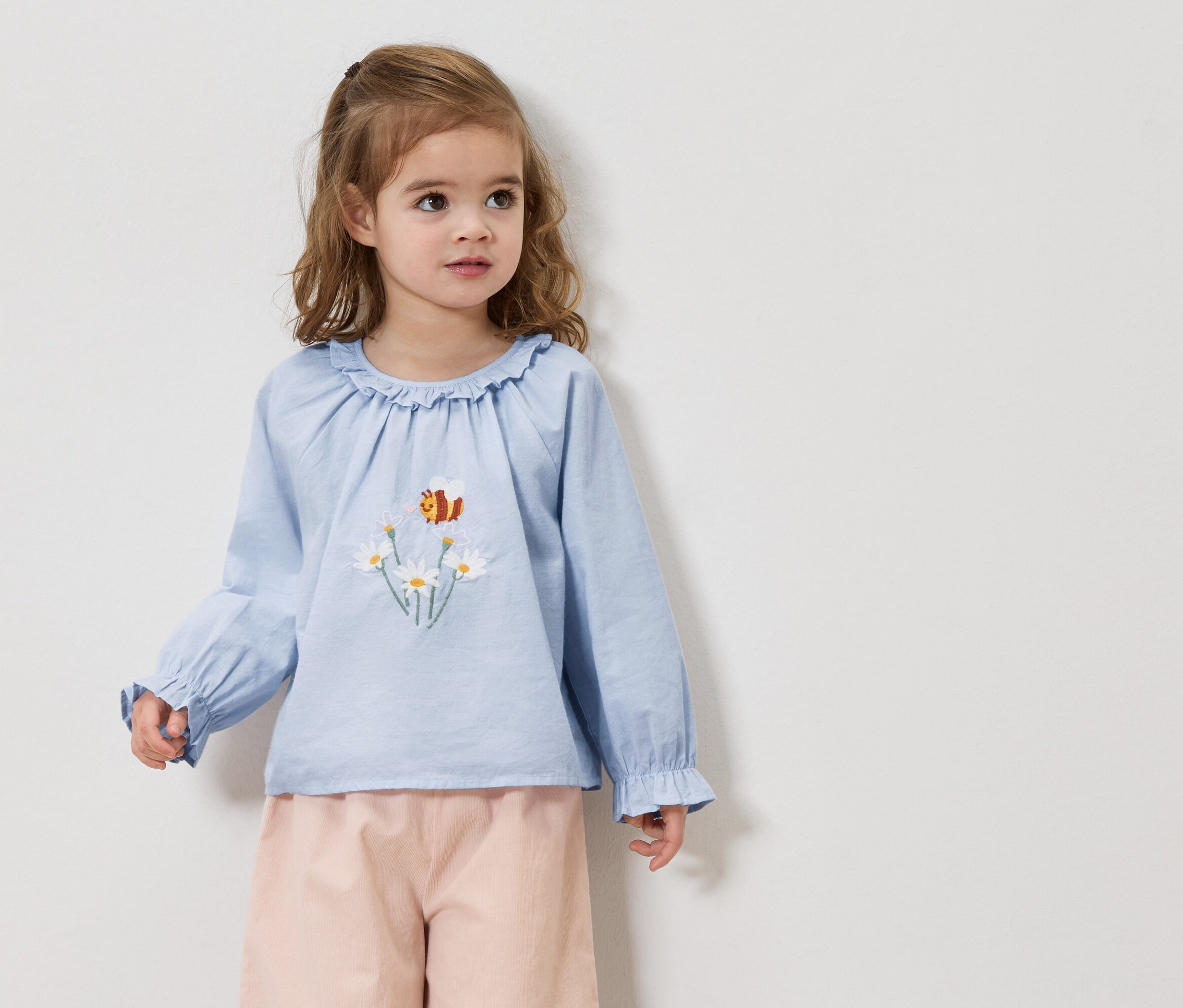 A szőke hajú kislány világoskék blúzt visel százszorszép és méh hímzéssel, valamint rózsaszín nadrágot.