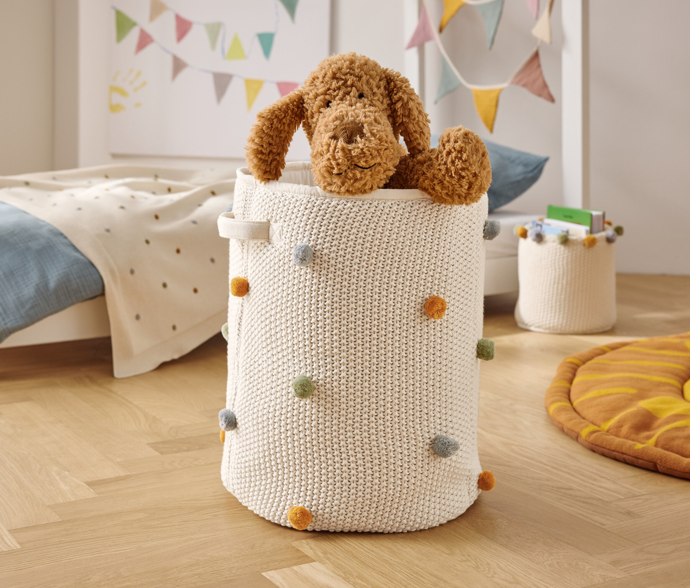 Egy barna plüsskutya ül egy fehér, kötött kosárban, színes pomponokkal.