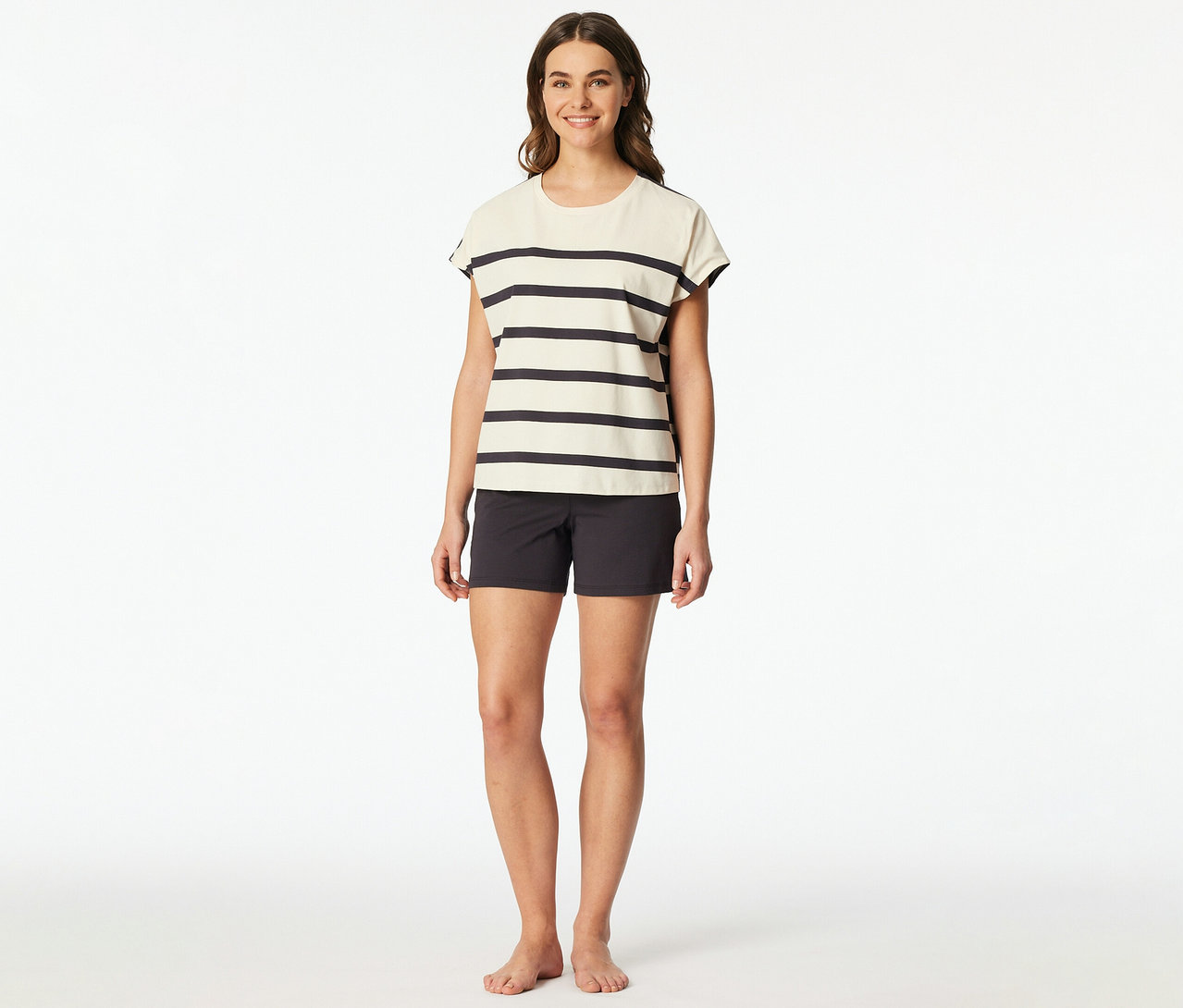 Mosolygó nő SCHIESSER rövidnadrágos pizsamát visel: krémszínű csíkos rövid ujjú felsőt és sötétszürke rövidnadrágot. Mezítláb van.