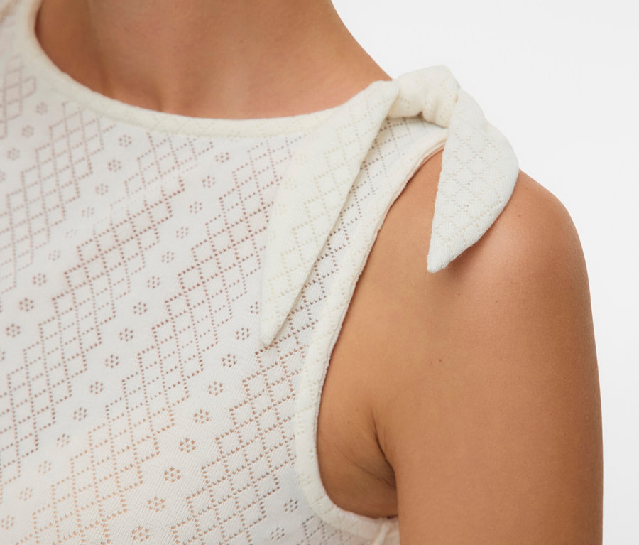 Egy nő közeli képe, aki egy ujjatlan, fehér, hímzett felsőt visel, aminek a vállán egy masni van.