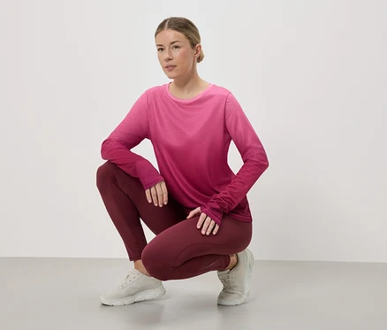 Guggoló nő sportruhában: rózsaszín-piros hosszú ujjú felső, bordó leggings és világosszürke tornacipő fehér háttér előtt.